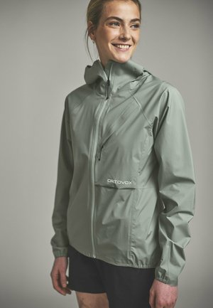 Lächelnde Frau trägt eine hellgrüne Ortovox Zip-Up-Jacke mit Kapuze und schwarzen Shorts vor neutralem Hintergrund.