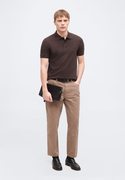 Jeune homme portant un polo marron foncé, un pantalon beige, une ceinture noire et des mocassins noirs, tenant une housse d'ordinateur portable noire d'une main, l'autre main dans la poche.