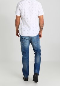 Camisa blanca de manga corta con pequeños patrones rojos y azules, combinada con jeans azul claro y zapatos negros. Vista desde la parte posterior.