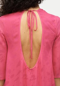 Ροζ φόρεμα με βαθιά πλάτη και κλείσιμο με κορδόνι. Το ύφασμα έχει μια διακριτική υφή με κηλίδες σε μοτίβο. Μανίκια κοντά.