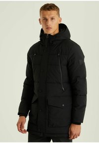 Schwarze Steppjacke mit Kapuze, ausgestattet mit einem Reißverschluss vorne, mehreren Reißverschlusstaschen und einer weichen, gesteppte Textur. Mit Druckknöpfen befestigt.