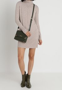 Femme portant une robe col roulé en maille beige, avec un sac bandoulière vert foncé, assortie à des bottines à talons vernies vert foncé.