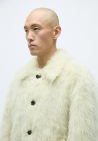 Pälsimitation i krämfärg med en strukturerad, fluffig yta. Har en stor krage och svarta knappar för stängning.