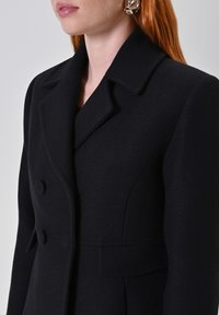 Blazer di lana nera con colletto a scollatura, frontale doppiopetto, finitura testurizzata e bottoni decorativi. Vestibilità su misura con cuciture strutturate.