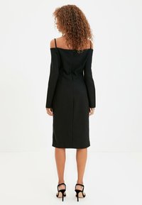 Černé midi šaty s odhalenými rameny a dlouhými rukávy, těsný střih a jemný zip na zadní straně. Na sobě má černé sandály na podpatku.