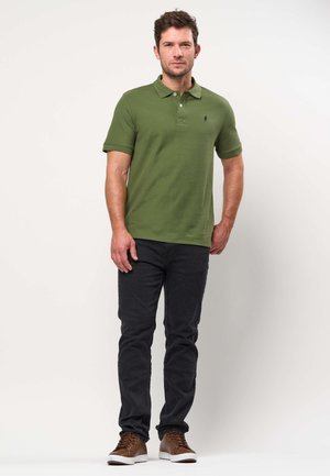 Férfi áll, olívazöld galléros pólót, fekete farmert és barna sportcipőt visel, egyszínű világos háttér előtt.