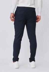 Pantalon slim bleu marine à texture lisse, avec deux poches arrière, associé à des baskets blanches sur un fond uni.
