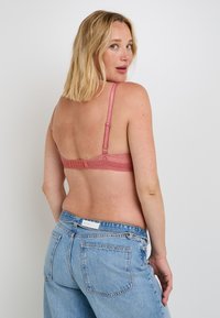 Bralette in pizzo rosa con spalline regolabili e chiusura a gancio. Modella che indossa jeans in denim blu sbiadito, mostrando il design sul retro del bralette.