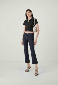 Woman wearing black cropped buttoned shirt, patterned navy blue pants, silver heeled sandals, and carrying small white shoulder bag.