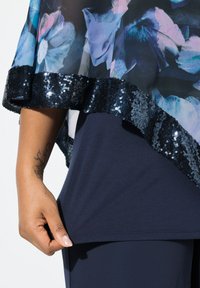 Cappa floreale trasparente con rifiniture in paillettes nere, sovrapposta a una maglia blu navy. Caratterizzata da un tessuto testurizzato e un design con orlo asimmetrico.