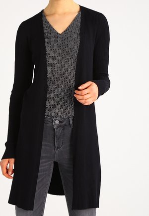 Frau trägt einen langen schwarzen gerippten Cardigan über einem schwarz-weiß gemusterten Top mit V-Ausschnitt und grauen Jeans.