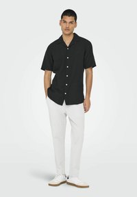 Camisa negra de manga corta con cuello y botones blancos, combinada con pantalones gris claro y zapatillas blancas, de pie sobre un fondo liso.