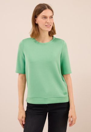 Femme portant un haut uni vert menthe à manches courtes et un pantalon noir, se tenant devant un fond neutre, regardant légèrement vers la droite.