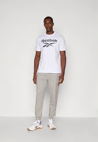 Biała bawełniana koszulka z ciemnoszarym logo "Reebok", zestawiona z jasnoszarymi zwężanymi spodniami dresowymi i białymi butami sportowymi z czarnymi akcentami.