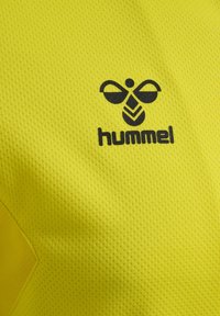 Żółty sportowy trykot wykonany z teksturowanej tkaniny, z czarnym logo pszczoły i napisem "hummel" na środku.