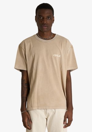 Giovane uomo con i capelli corti che indossa una t-shirt beige con il logo "CYCLE" e pantaloni chiari, in piedi davanti a uno sfondo bianco semplice.