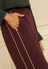 Burgundy-byxor med smal passform, sidofickor och en guldkant längs den yttre sömmen, tillverkade av ett mjukt tyg.