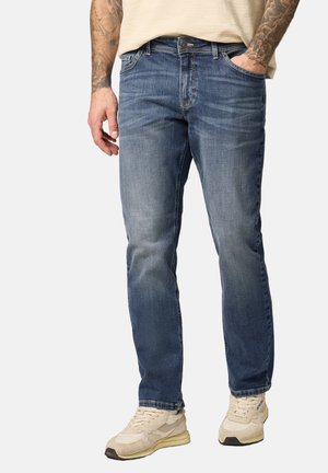 Mann mit tätowierten Armen, der verblasste blaue Jeans und cremeweiße Sneakers trägt und mit einer Hand in der Tasche vor einem einfarbigen Hintergrund steht.