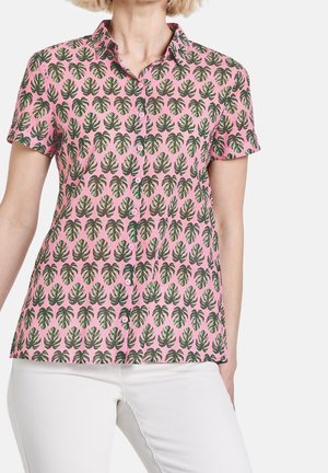 Short-sleeve pink button-up shirt with green palm leaf pattern worn with white pants, shown from neck to hips.