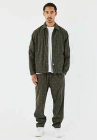 Chaqueta verde oliva y pantalones a juego, hechos de un tejido suave y ligero, con un sutil patrón repetido y cierre de botones.