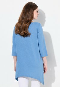 Hellblaues Tunika mit Dreiviertelärmeln, abgerundetem Saum und weichem Griff, kombiniert mit weißen Hosen. Die Rückansicht zeigt Haare, die über die Schulter fallen.