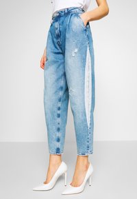 Jeans in denim di colore azzurro chiaro con dettagli strappati, vita alta, gamba affusolata e striscia bianca laterale. Indossati con tacchi a punta bianchi.