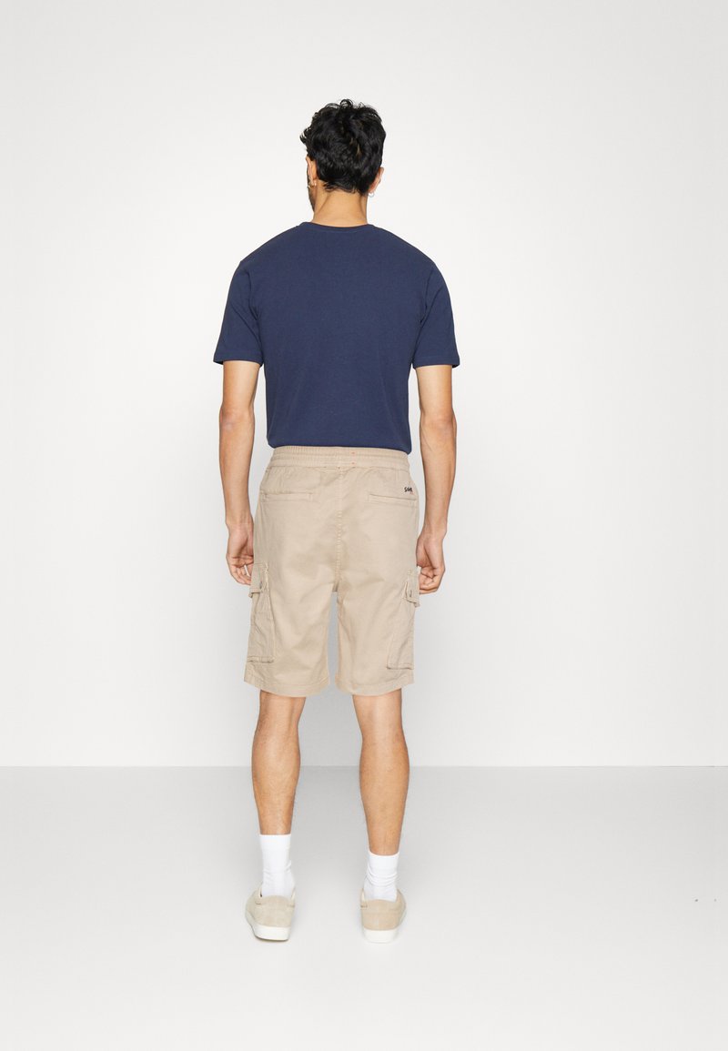T-shirt à manches courtes bleu marine, short cargo beige clair avec poches latérales, tous deux en coupe décontractée. Le modèle porte des chaussettes blanches et des chaussures claires.
