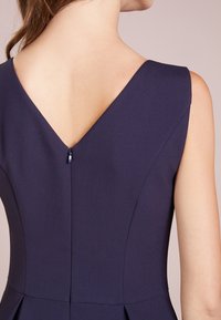 Marineblaues, figurbetontes Kleid mit V-Ausschnitt, ärmellosem Design und einem verdeckten Reißverschluss an der Rückseite, gefertigt aus glattem Stoff.