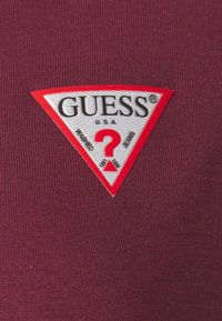 Maroon kangas yenye nembo ya pembetatu ya rangi nyekundu na nyeupe ya "GUESS U.S.A." ikiwa na maandiko ya "WASHED" na "JEANS" na kipengele cha alama ya swali katikati.