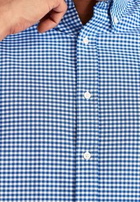 Camisa a cuadros azul y blanca con cuello abotonado, presenta pequeños botones y una textura de tela suave.