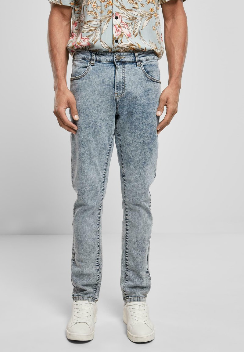 Jean en denim délavé avec une coupe slim, présentant un design à cinq poches, des détails cousus et une texture légèrement passée. Porté avec des baskets blanches.
