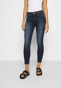 Jeans ajustados de color azul oscuro con cintura alta, que cuentan con cierre de cremallera, bolsillos frontales y bajo deshilachado. Combinados con sandalias planas negras.