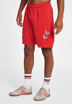 Atletisk person iført røde sportsshorts med hvid Chevron-logo, hvide sokker med sorte og røde striber og beige sneakers med orange chevroner.
