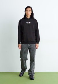Sort hættetrøje med BMW-logo og "Motorsport" tekst, parret med grå, teksturerede jeans og sorte sneakers. Simpel, afslappet påklædning.