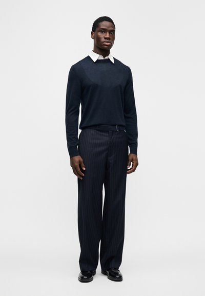 Μπλε πουλόβερ με λευκό γιακά, σε συνδυασμό με μπλε παντελόνι με λευκές ρίγες. Το μοντέλο φοράει μαύρα γυαλιστερά παπούτσια. Απλός και κομψός σχεδιασμός.