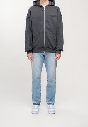 Osoba ubrana w ciemnoszary rozpinany kaptur z przednimi kieszeniami, jasnoniebieskie dżinsy i białe oraz jasnoniebieskie trampki, stojąca na jednolitym tle.