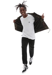 Vit t-shirt, svarta joggers och svarta högskor. Camo-fodrad jacka hängande öppen, med dragkedja och logotypdetaljer.