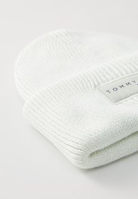 Bonnet en tricot blanc avec un revers, comportant une étiquette rectangulaire avec "TOMMY" en texte bleu foncé sur un fond clair. Texture lisse.