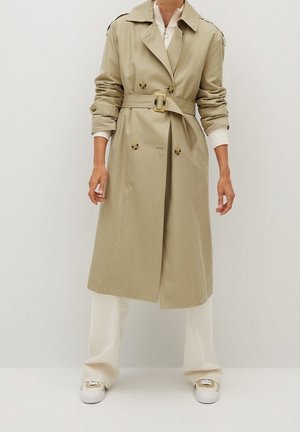 Persoon die een beige trenchcoat met riem draagt, witte wijde broek en beige sneakers, staand tegen een effen lichte achtergrond.