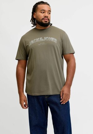 Mand med flettet hår og skæg, iført en olivengrøn Jack & Jones t-shirt og blå jeans, kigger sidelæns mod en ensfarvet baggrund.