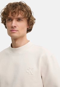 Felpa color crema chiaro in morbido materiale, caratterizzata da un collo alto e un logo cK goffrato sul petto. Design casual con una vestibilità comoda.
