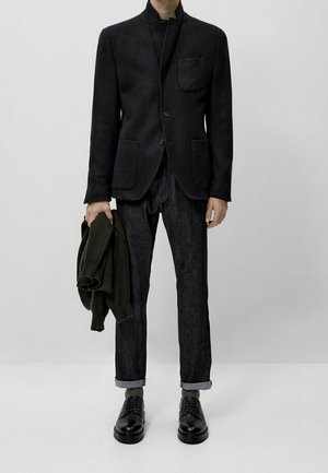 Svart kavaj med mandarin-krage och ficka, mörka jeans med uppvikta fållar, grön tröja och svarta snörskor på en neutral bakgrund.