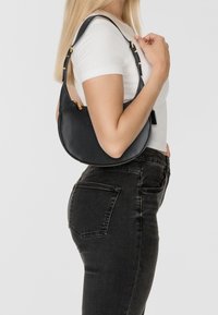 Borsa a spalla in pelle nera con forma curva, dettagli in metallo dorato, tracolla regolabile e chiusura con zip. Indossata in modo casual con una maglietta bianca.