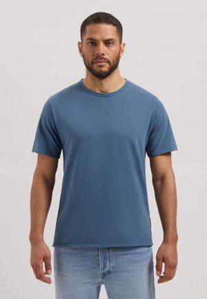 Man met kort haar en baard, gekleed in een effen blauw T-shirt met korte mouwen en lichtblauwe spijkerbroek, staand tegen een neutrale achtergrond.