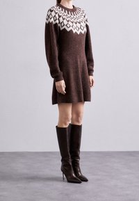 Robe en tricot marron avec un empiècement en losanges blancs, des manches longues et un ourlet évasé ; associée à des bottes noires à talons montantes.