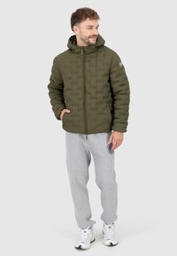 Olijfgroene pufferjack met capuchon, voorzien van een gestikt patroon en ritssluiting, gecombineerd met grijze sweatpants en witte sneakers.