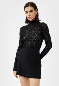 Zwarte kanten longsleeve-top met hoge hals en ingewikkeld bloemmotief, gecombineerd met een aansluitende zwarte minirok met een split aan de voorkant.
