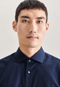 Granatowa koszula zapinana na guziki z wyraźnym kołnierzykiem, gładką fakturą i dwoma jasnymi guzikami z przodu, noszona przez osobę zwróconą przodem.