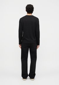 Άνδρας με σγουρά μαλλιά, φορώντας μαύρο μακρυμάνικο πουκάμισο, μαύρο τζιν και λευκά παπούτσια, στέκεται γυρισμένος με την πλάτη προς ένα απλό φόντο.