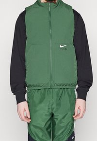 Man i grön ärmlös Nike Air zip-up väst över svart långärmad tröja och matchande gröna träningsbyxor med vita ränder.